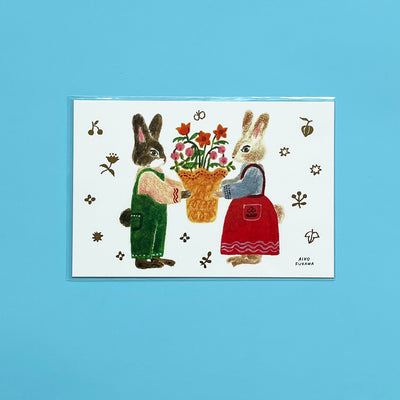 A postcard with an illustration of two anthropomorphic rabbits in outfits holding a bouquet of flowers on a white background.