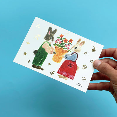A hand holding the postcard with rabbits at an angle to show the shimmer of the gold floral details.