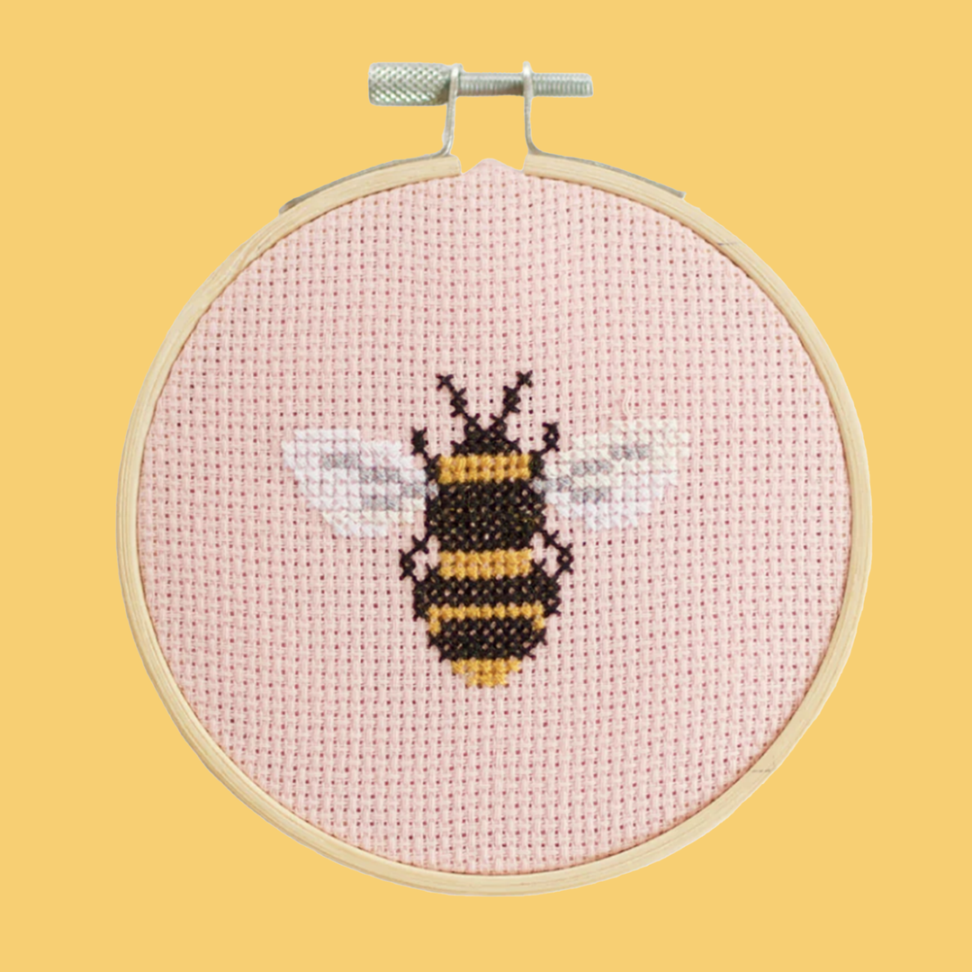 A finished cross stitch project featuring a bumble bee with out stretched wings. The project is diplayed in an embroidery hoop on a yellow background.