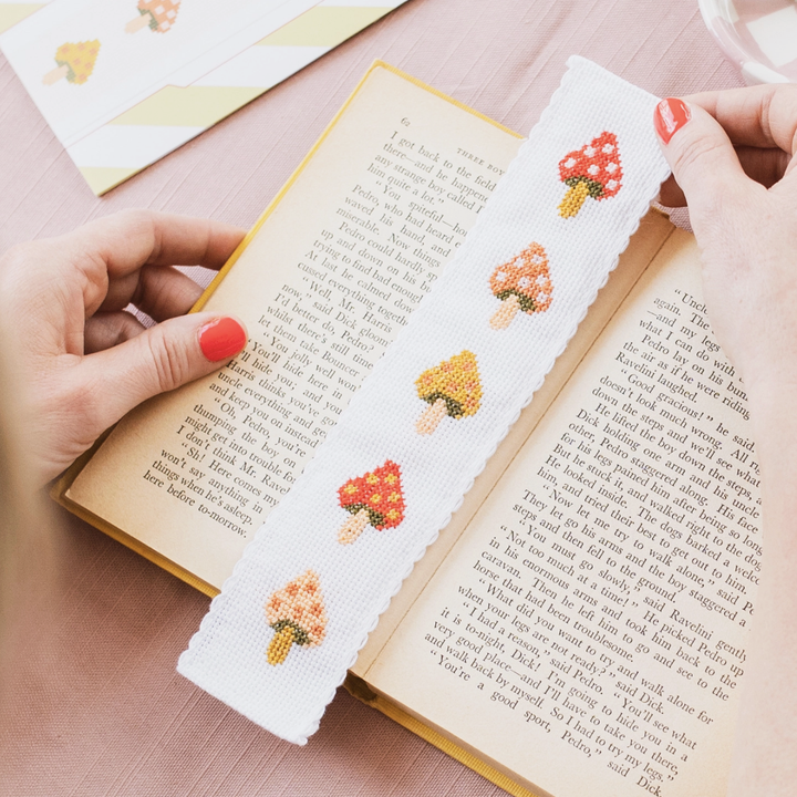 Hands holding a fabric book mark on top of an open book. The book mark features five mushrooms cross stitched on it.