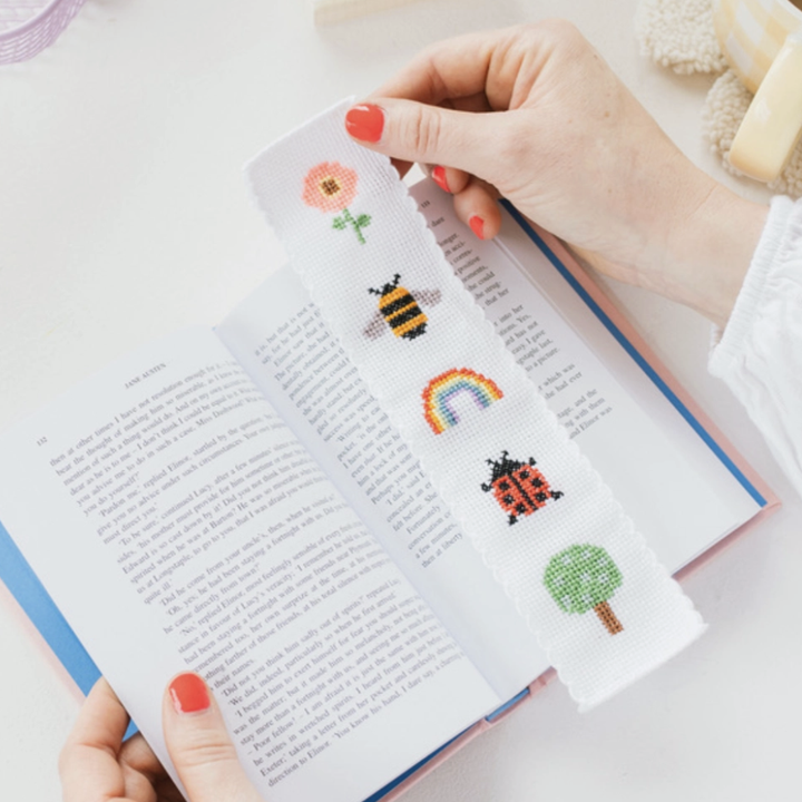 Hands holding a fabric book mark on top of an open book. The book mark features a cross stitch design of a flower, bee, rainbow, lady bug and tree.