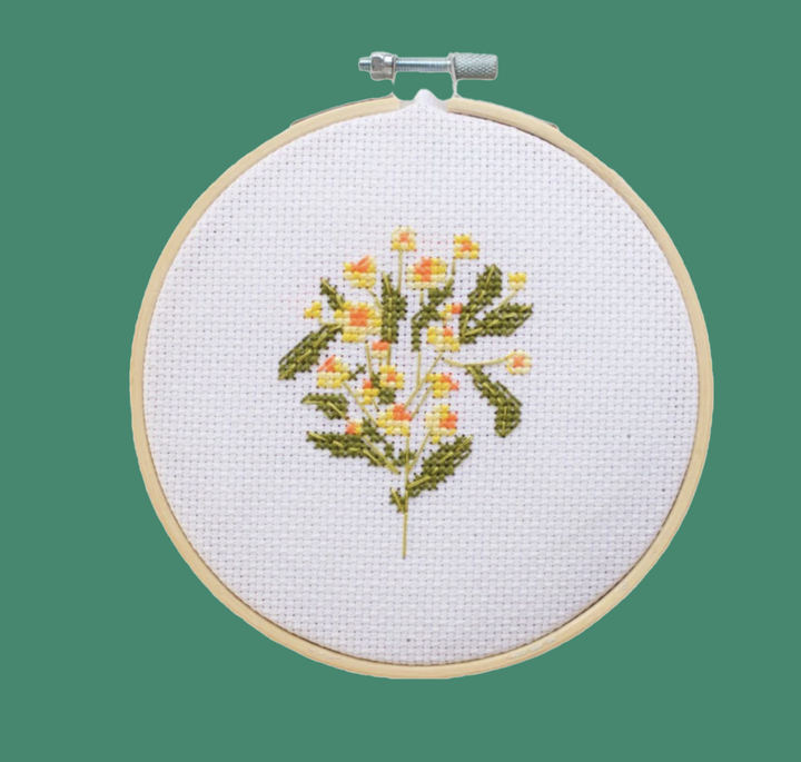 A finished cross stitch project featuring a sprig of chamomile flowers displayed in an embroidery hoop on a bright green background.