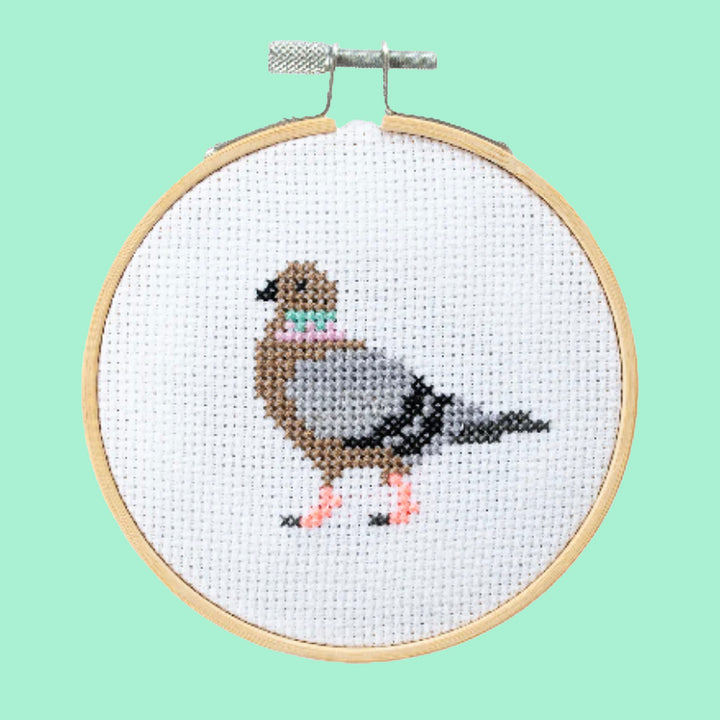 A finished cross stitch project featuring a grey pigeon in profile. The project is displayed in an embroidery hoop on a light green bacground.