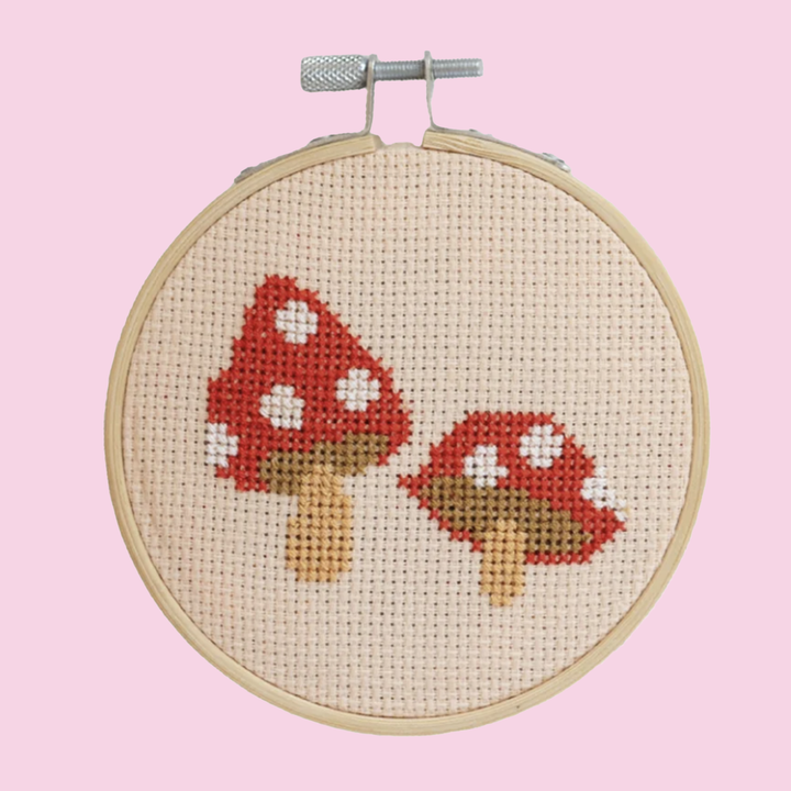 A finished cross stitch project featuring two red mushrooms with white spots. The project is displayed in an embroidery hoop on a bright pink background.