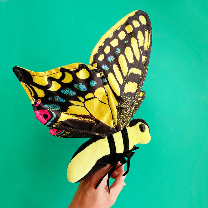 An oversized yellow and black swallowtail finger puppet with butterfly wings, held by a hand against a green background.