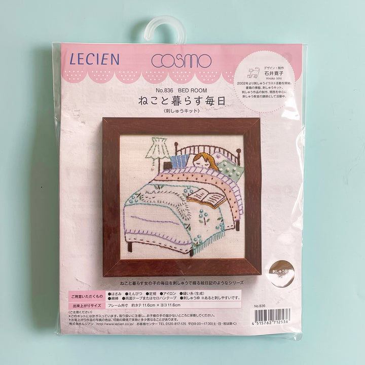 Packaged embroidery kit featuring a girl laying in bed covered with colorful blankets. A fluffy white cat sleeps at the foot of the bed.