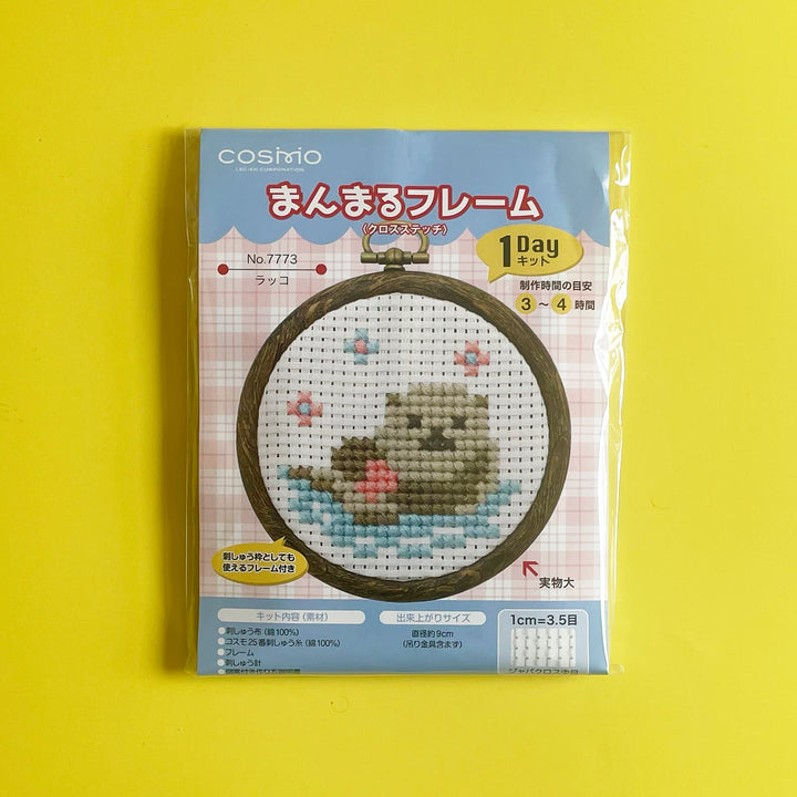 Packaged cross stitch kit featuring a light brown otter floating in the water with a pink shell in its lap.