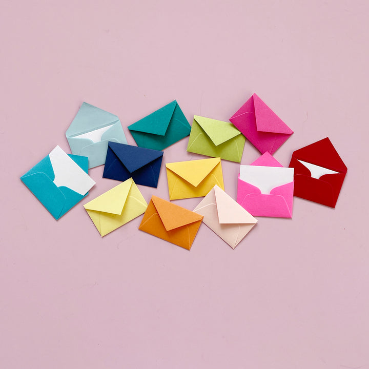 Twelve miniature envelopes in a rainbow of  colors artfully scattered on a light pink background.