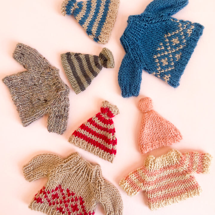 Four miniature, knit sweaters with matching hats, artfully scattered on a light pink background.