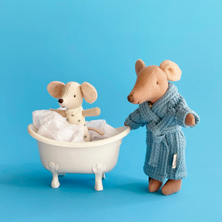 Two miniature fabric mice set up in a bath scene. A smaller mouse plays in a white metal bathtub, while a larger mouse in a blue, waffle weave bathrobe supervises.