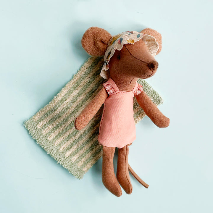 A miniature fabric toy mouse dressed in a pink swimsuit and floral sun hat laying on a small striped towel on a light blue background.