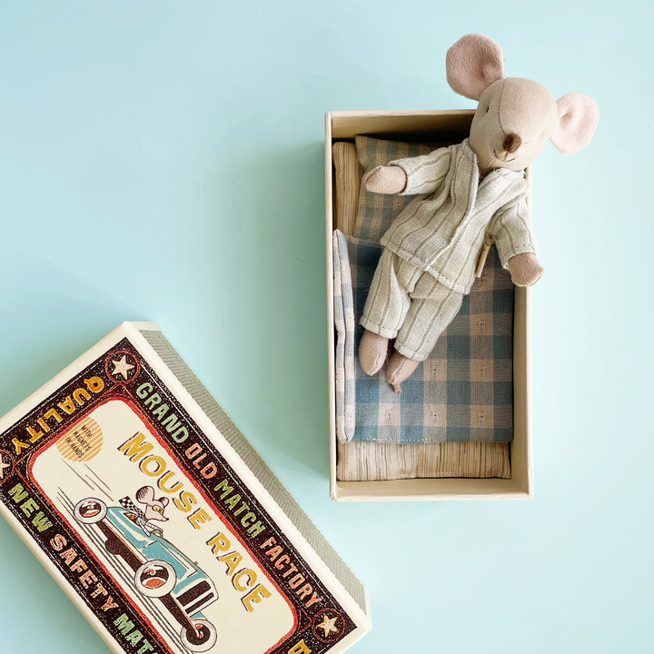 A miniature toy fabric mouse tucked into an open, oversized matchbox . the mouse wears a pair of striped pajamas and sit on top of a blue gingham blanket.