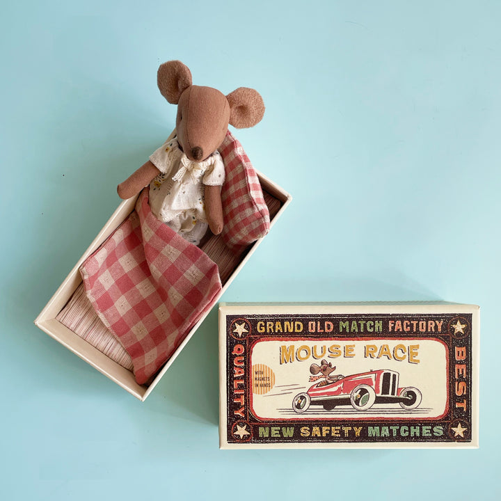A miniature toy fabric mouse sitting in an open, oversized matchbox. The mouse is wearing a pair of cream pajamas and is covered by a red gingham blanket.