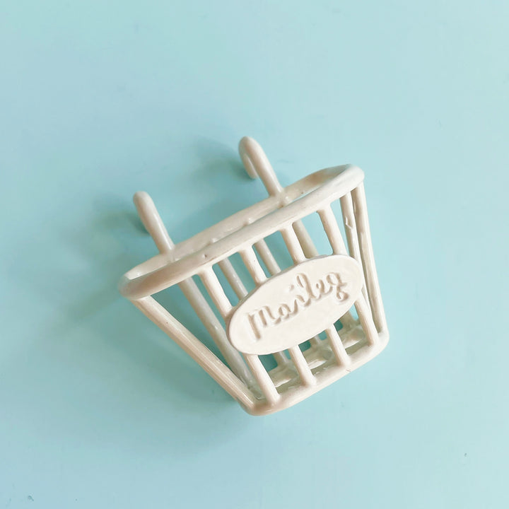 Small, white metal basket with loops branded with a Maileg stamp on a light blue background.