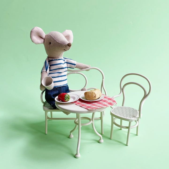 Stuffed mouse in a striped shirt sitting at a miniature table with food on a light green background