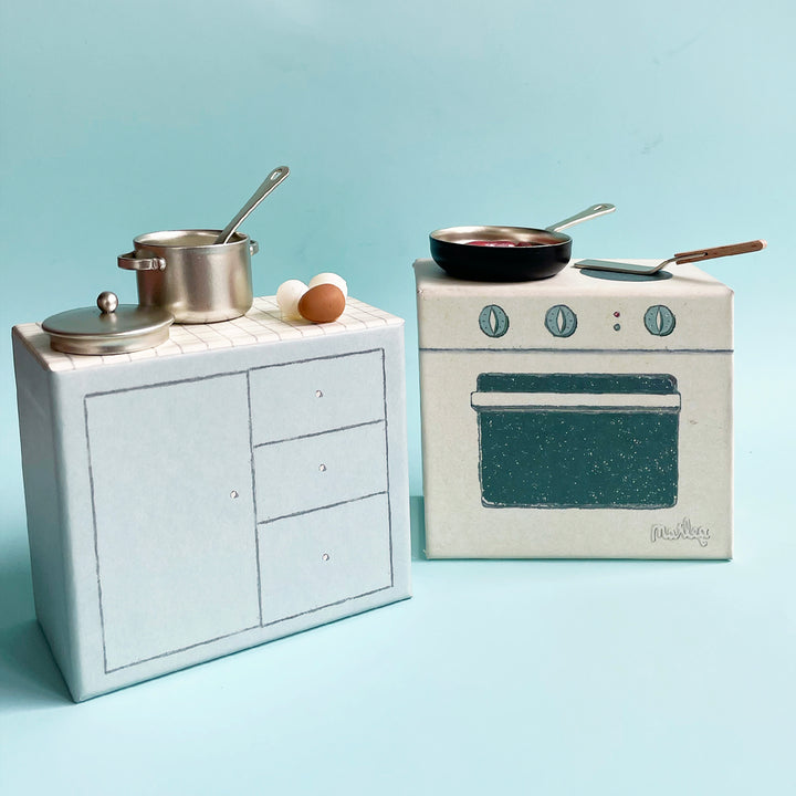 Miniature, illustrated paper-board kitchen set displayed with metal cookware, utensils and tiny toy eggs on a light blue background.