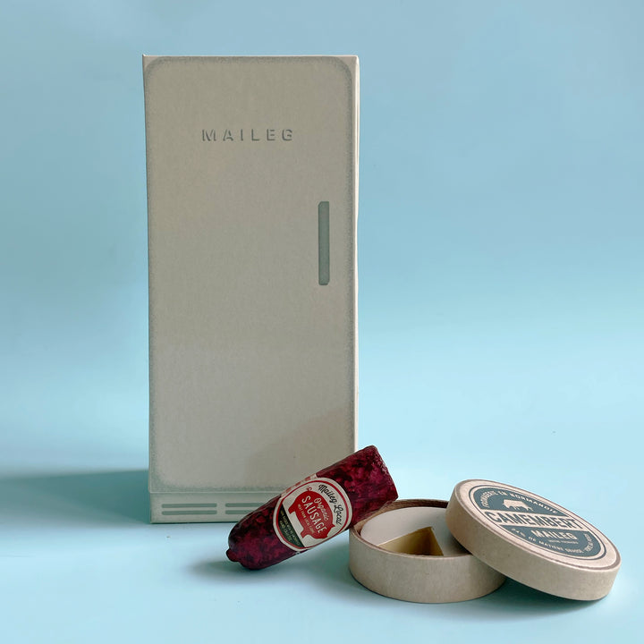 Miniature Maileg paper-board fridge with tiny toy sausage and cheese displayed on a light blue background.