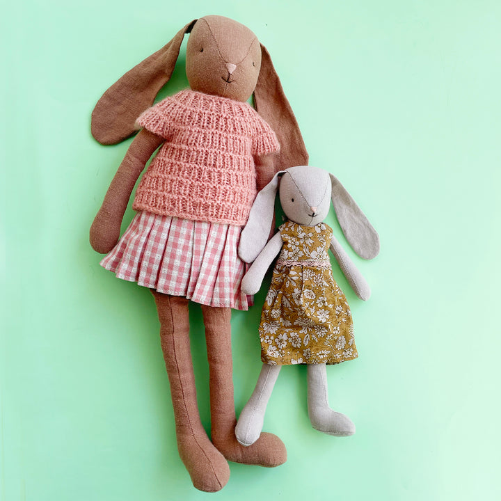 Two stuffed cotton rabbits: one large; one small. The large rabbit wears a pink, knit sweater and a pink gingham skit. The small rabbit wears a floral dress.