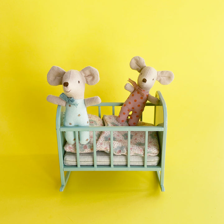 Two miniature fabric baby mice play in a mint-colored metal cradle with floral bedding against a bright yellow background.