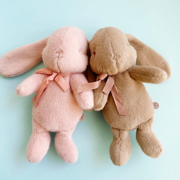 Two fuzzy, stuffed rabbits with long floppy ears lay side by side against a light blue background. One rabbit is pink, the other is brown. they both wear a silk bow around theitr necks.