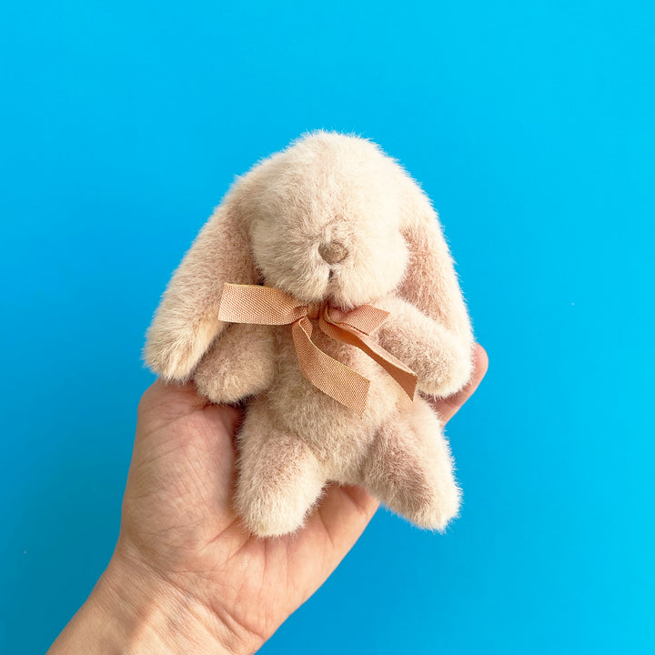 A small, fuzzy, light pink rabbit with floppy ears and a silk neck bow is held in the palm of a hand against a brigt blue background.
