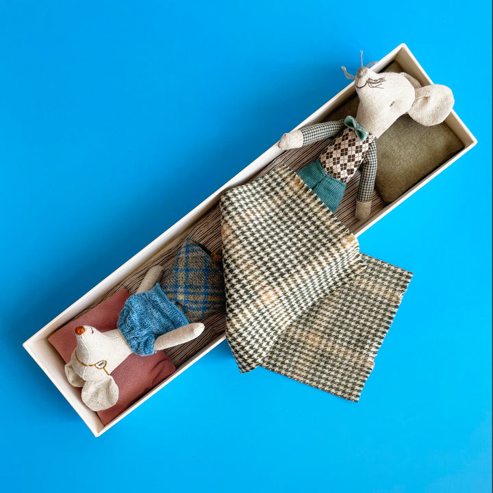 Two miniature fabric toy mice are nestled into a long, oversized matchbox. One mouse sits on each end, covered by a plaid woolen blanket.