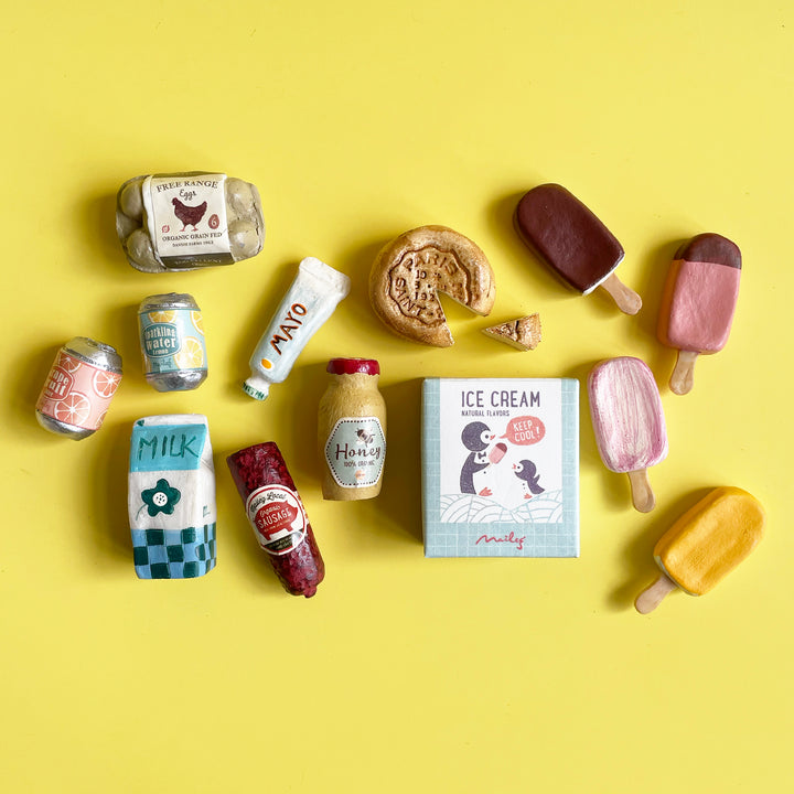 A collection of miniature, hand-painted play food, including soda cans, sausage, milk and ice cream artfully scattered on a bright yellow background.