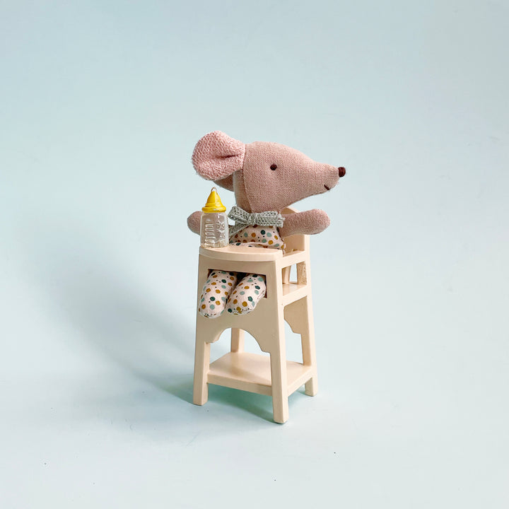 A miniature fabric baby mouse sits in a white wood highchair with a tiny plastic baby bottle sitting on the tray, againsta  light blue background.