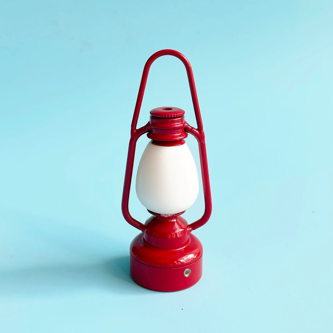 A miniature, red metal, vintage inspired lantern displayed against a light blue background.