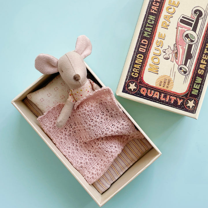 A miniature fabric toy mouse tucked into an open, oversized matchbox. The mouse lays on a striped mattress and is covered by a pink knitted blanket.