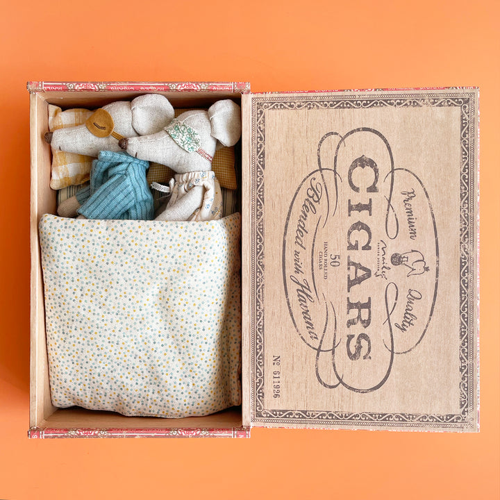 Two miniature fabric toy mice lay side by side, snuggled into an open cigar box. Both mice wear eye masks and are covered with a padded, polka-dot blanket.
