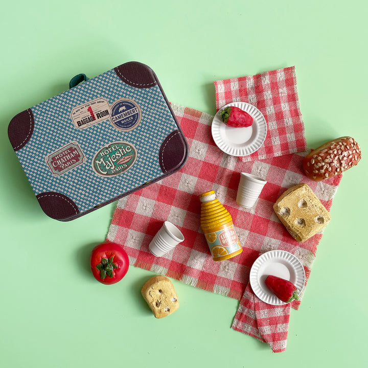 Miniature toy picnic artfully scattered on a light green background. Red checkered table cloth is laid out with tiny plates and tiny, hand-painted food items, including cheese, tomato and bread.
