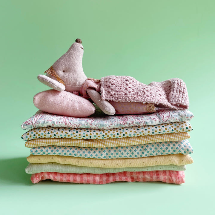 A miniature fabric mouse laying on top of a stack of 7 fabric mats, with pink pillow and pink knit blanket. Displayed on a light green background.