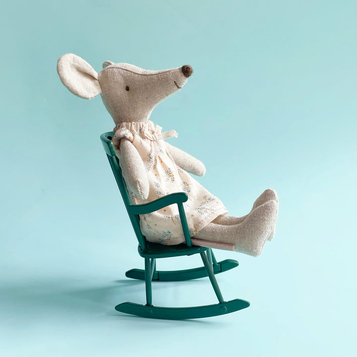 A Maileg fabric mouse in a floral dress sitting in a miniature, dark green metal rocking chair against a light blue background.