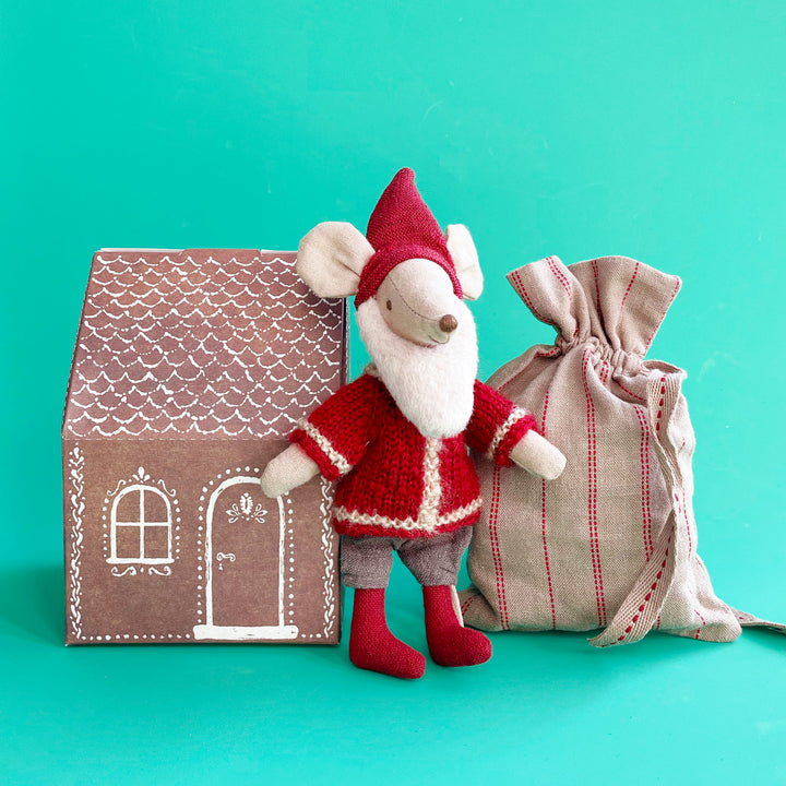 A miniature fabric mouse dressed in a knitted Santa suit, and fluffy beard. The mouse stands between a small paper house decorated to look like a gingerbread house, and a red-striped burlap sack.
