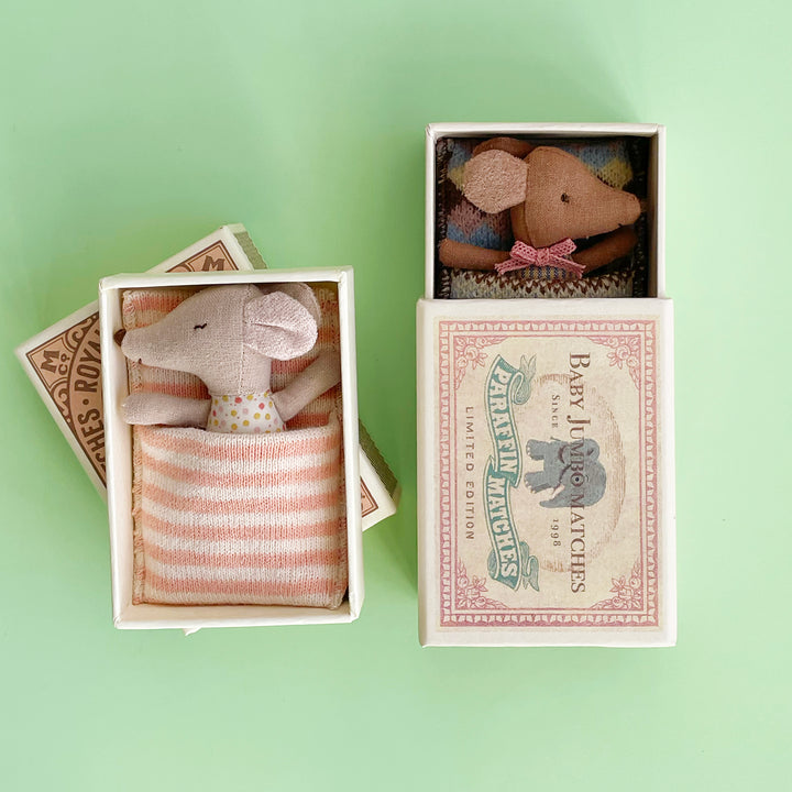 Two miniature fabric mice tucked into colorful matchboxes.One baby mouse has its eyes open and one has its eyes closed.