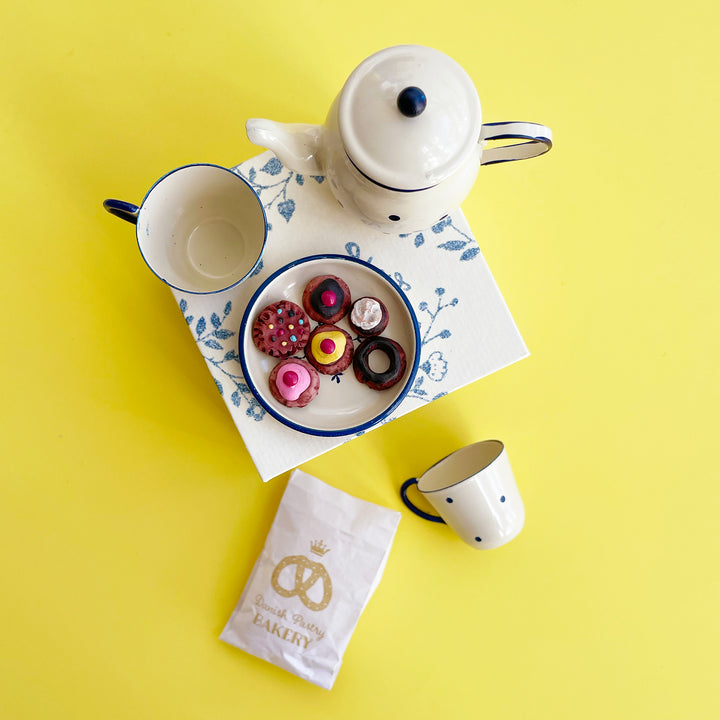 An overhead photo of a miniature metal tea set with navy blue details sitting on top of a blue floral box against a bright yellow background. A plate of tiny toy cookies sits next to the teapot.