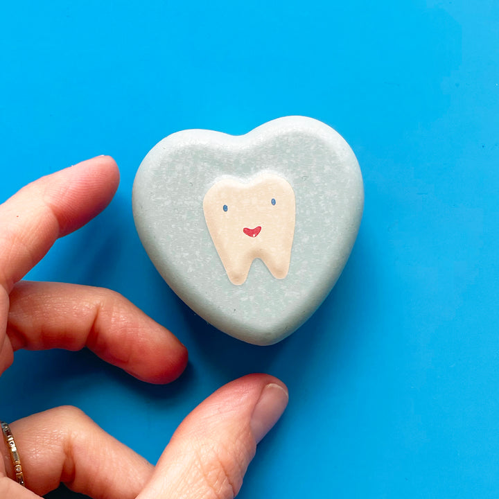 A hand reaches in from the left to touch a tiny heart-shaped tin made of mint-colored metal with an embossed white tooth with a smiley face on it.