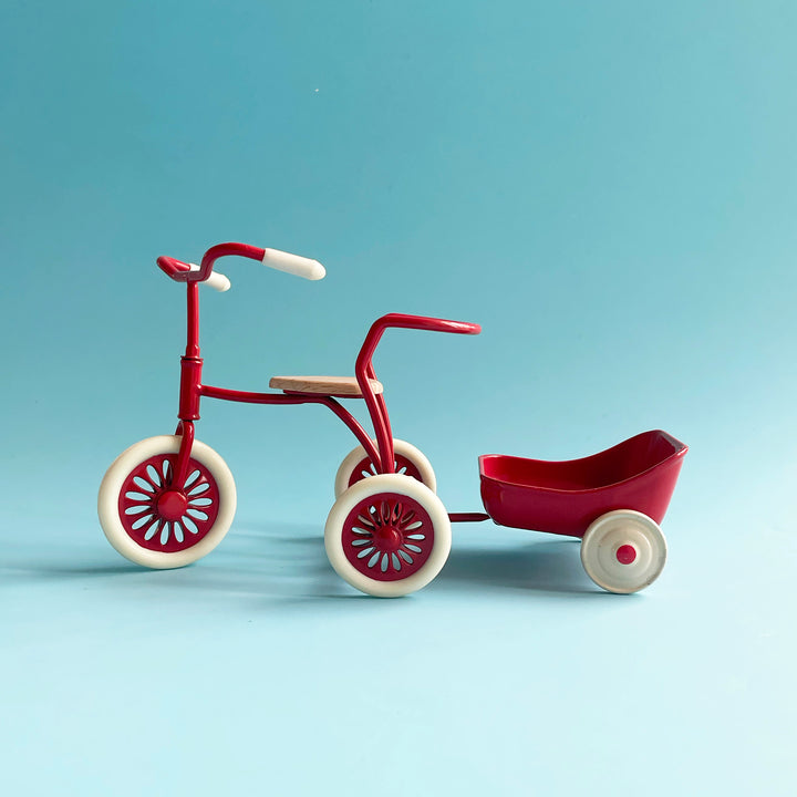 A miniature, red metal tricycle with white handlebars and wheels pulls a small, red metal cart against a light blue background.