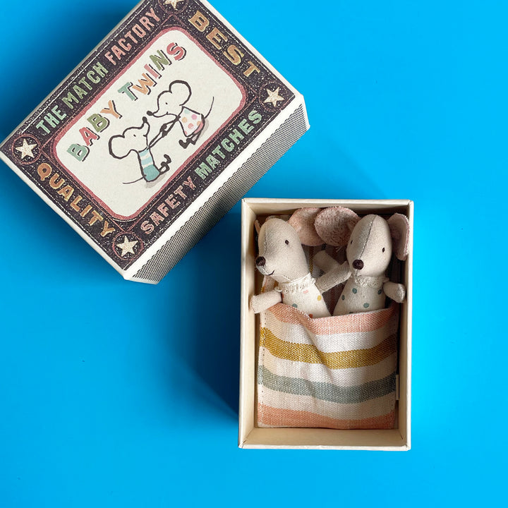 Two miniature fabric baby mice snuggled together under a striped blanket in an open, oversized matchbox against a blue background.