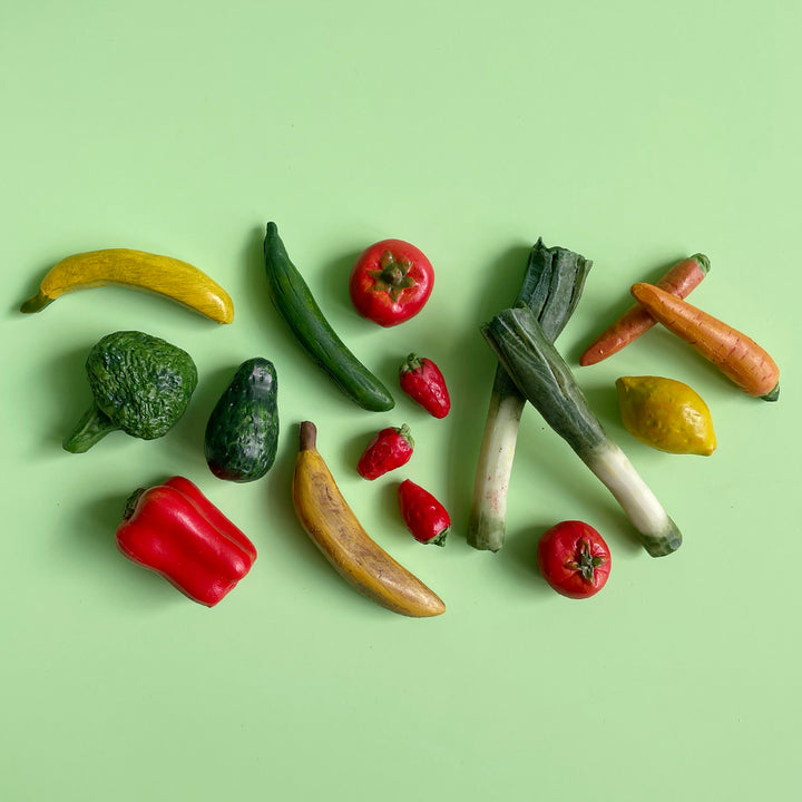 Miniature hand-painted toy vegetables, including bananas, tomatoes, leeks and strawberries, artfully scattered on a light green background.