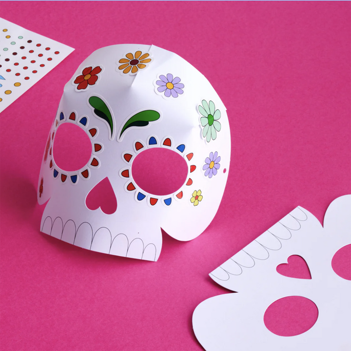 A DIY Calavera Mask decoratd with Stickers on a bright pink background.