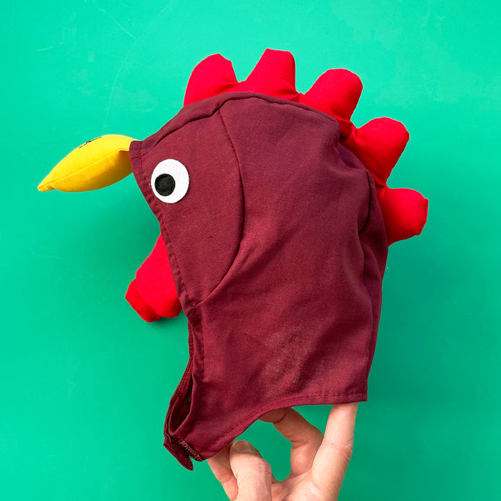 Maroon cotton chicken costume hat with yellow padded beak, red padded crest and felt yes. Displayed on a bright green background.