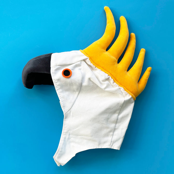 White cotton cckatoo costume hat with black cotton-velour beak, yellow cotton-velour stuffed crest and felt eyes. Displayed on a bright blue background.
