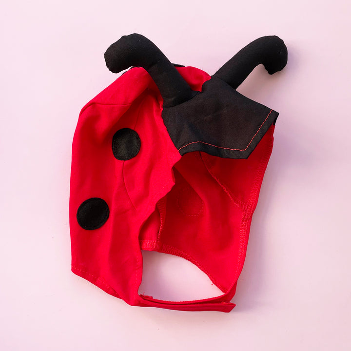 Red cotton ladybug costume hat with black padded antennae and felt spots. Displayed on a pink background.