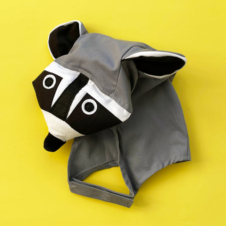 Grey cotton raccoon costume hat with a stuffed snout and felt accents. Displayed on a bright yellow background.