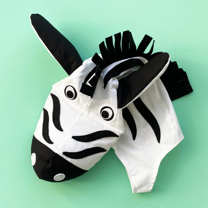 Wite cotton zebra costume hat with black felt stripes, a padded snout, floppy ears and a black felt mane. Displayed on a light green background.