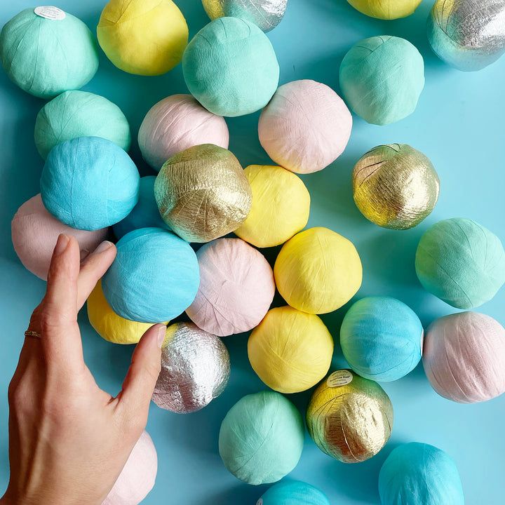 A pile of small pastel and metallic surprise balls on a light blue background with a hand reaching in to grab one.