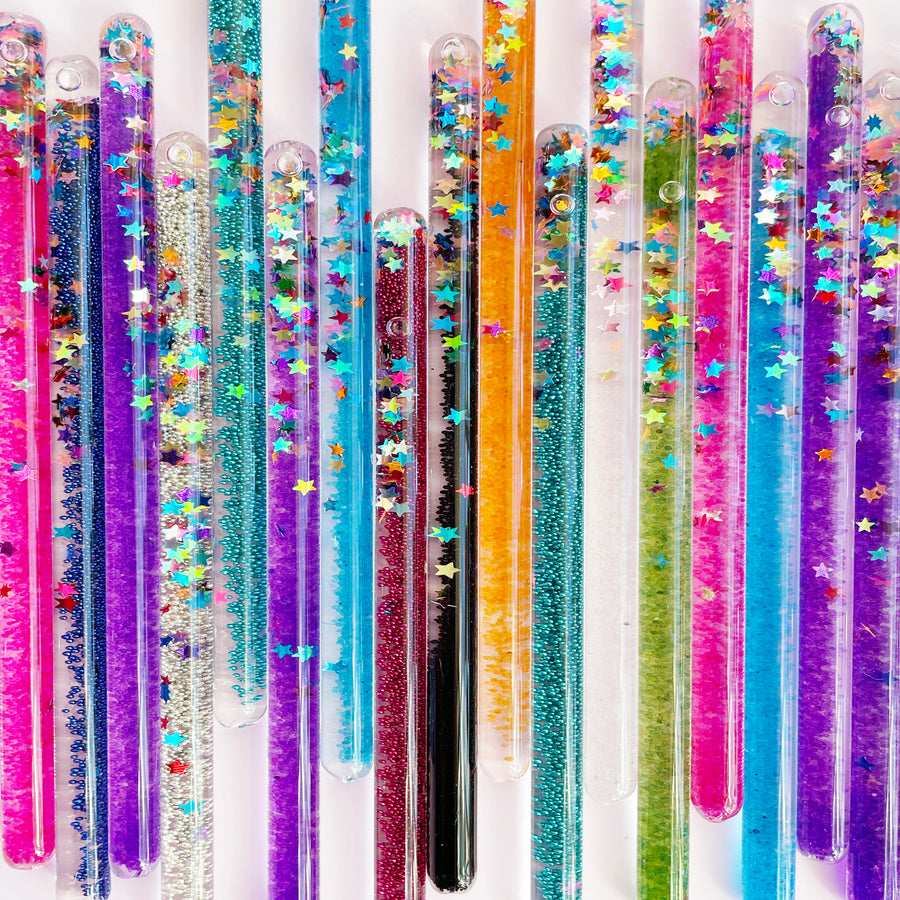 A close-up shot of a row of clear plastic wands filled with colorful glitter and sparkling stars on a light pink background.