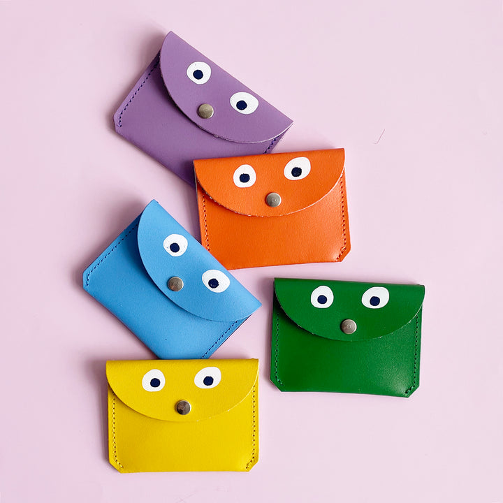 Several colorful googly-eye coin purses with metal button on light pink background.